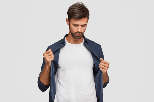 People, clothing and advertisement concept. Confident unshaven young Caucasian man dressed in fashionable shirt, shows blank copy space on white t shirt for your slogan or promotional content.