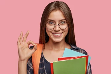 Fotobehang Cheerful pupil being in good mood after passing exam, shows with gesture everything is okay or excellent, holds colourful folders, dressed in shirt, has rucksack, isolated over pink background © Wayhome Studio