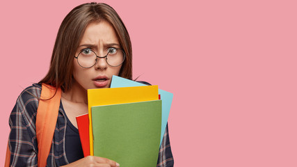 Fototapeta premium Horizontal shot of scared brunette woman with puzzled expression, frowns face, holds many books, feels nervous before answering during class, stands against pink studio wall with blank space