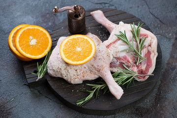 Black wooden chopping board with raw duck legs, fresh rosemary and orange slices, grey stone background, studio shot