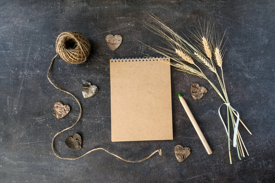Top view of craft Recycle paper notebook with bunch of wheat spikes, twine, wooden pencil and bark hearts on dark table. Recycle, natural, rustic and eco mockup background with copy space. Flat lay