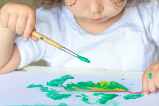 Summer or Autumn.Close up of hand little kidl drawing with green paint on fall lives. Curly adorable child toddler painting at table.