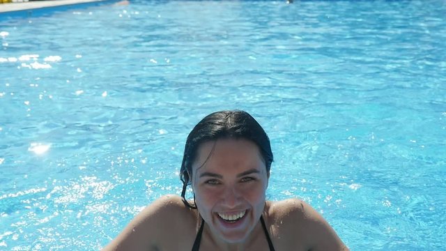 An exciting view of a smiling young woman in black bikini jumping hilariously at the edge of a pool with sparkling celester waters in slo-mo