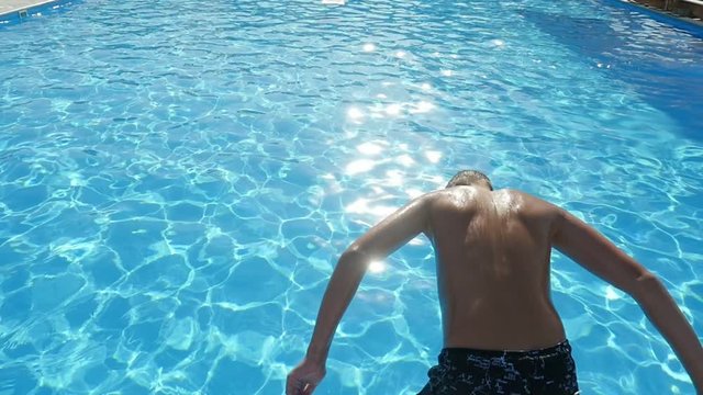 An exciting view of a 12 year old boy in long shorts jumping in the turquiose water of the modern swimming pool in summer in slow motion