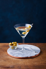 Martini cocktail with green olives on marble board. Wooden table. Blue background.