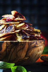 Chips from purple and red potatoes in wooden bowl on background of vegetables, selective focus