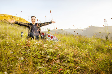 A young traveler sits with hands in a sides on a hill in the background of a mountain.