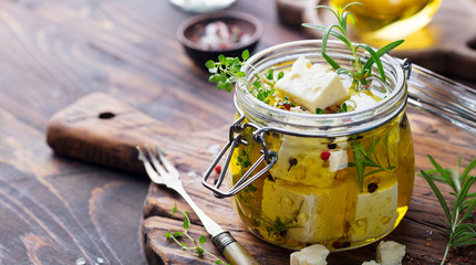 Feta cheese marinated in olive oil with fresh herbs in glass jar. Wooden background. Copy space. - obrazy, fototapety, plakaty