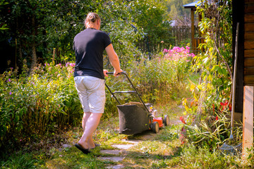 Gardener with electric lawn mower is trimming the garden. Sunny day, blooming garden. Adult man...