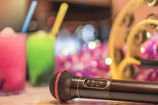 Black microphone in karaoke club, with remote controller, melon and strawberry soda drinks, yellow tambourine and screen for singing music on stage party.