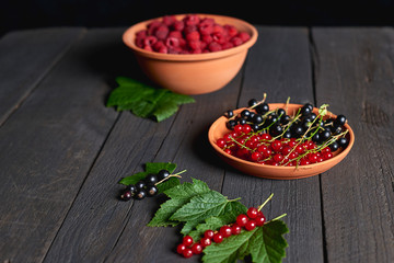 Ceramic plate with black and red currant and bowl of red raspberries on black rough wooden texture table