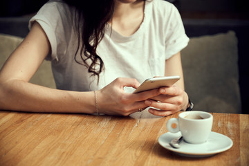 Close up women's hands holding smartphone. Young woman sitting in coffee shop at wooden table, drinking coffee and using smartphone. Female browsing internet, chatting, watching video on mobile phone.