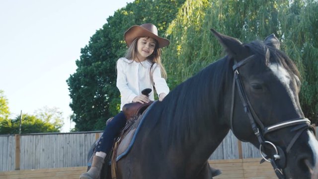 Pretty little girl in hat riding a black mare on the arena. 4K