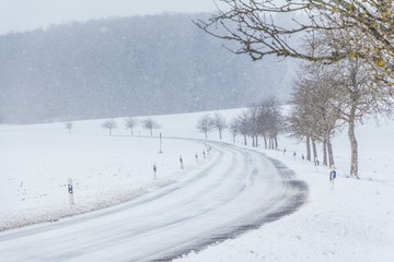 Fototapeta premium Leere glatte Winter Straße mit Schnee wartet auf Winterdienst. Die Fahrbahn ist weiß glatt verschneit. Vorsicht vor Unfall durch Schneesturm und schlechte Sicht. Wetter zur Weihnachten Jahreszeit