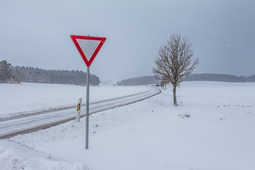 Leere glatte Winter Straße mit Schnee und Vorfahrt achten Schild. Die Fahrbahn ist weiß glatt verschneit. Vorsicht vor Unfall durch Schneesturm und schlechte Sicht. Wetter zur Weihnachten Jahreszeit