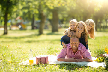 A cheerful family sitting on the grass during a picnic in the park, there is a basket with food and...