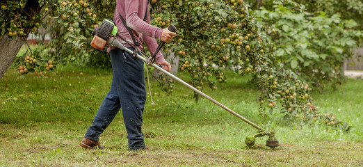 Gardener using machine cutting green grass in garden. Garden equipment. Young man mowing the grass with a trimmer