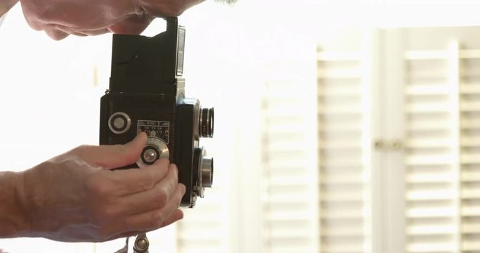 A Caucasian male photographer standing in front of a bright sunlit window with shutters looks into the viewfinder of a vintage TLR style camera.
