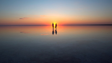A young couple walking on calm mirrored surface of shallow water into the setting sun.. They are...