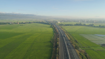 Aerial view highway with cars among farmer fields, rice terraces on background of mountains. Philippines, Luzon. High speed highwayin the morning sunrise. Tropical landscape in Asia.