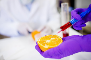Close up of a hand in a protective glove holding orange and taking a sample from it with a syringe.