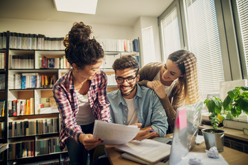 Two cheerful student girls helping their friend to pass the exam.Sitting in a bright library.