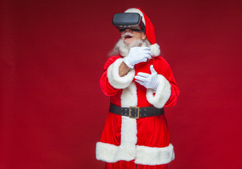 Christmas. Santa Claus in black virtual reality glasses holding a red bucket of popcorn. Watching a movie in virtual reality. Isolated on red background.