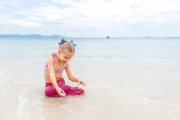 Little beautiful girl dressed in swimsuit as a mermaid sits on the seashore