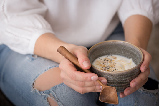 Closeup of bowl with homemade organic vanilla yogurt with added chia seeds and coconut chips in hands of a young woman sitting on a sofa
