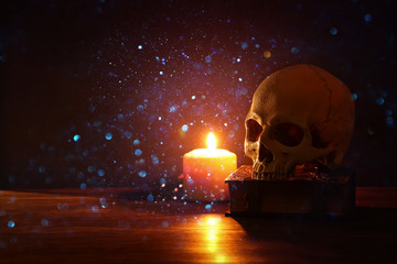 Human skull, old book and burning candle over old wooden table and darl background.