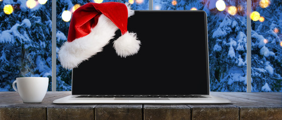 Laptop on table with Santa Claus hat at home with panoramic view through window of snowy trees in winter forest, screen is cut with provided clipping path