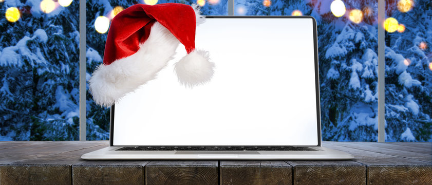 Laptop on table with Santa Claus hat at home with panoramic view through window of snowy trees in winter forest, screen is cut with provided clipping path
