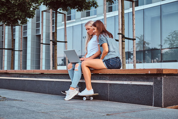 Two beautiful hipster girls sitting on the bench with a laptop on a background of the skyscraper.
