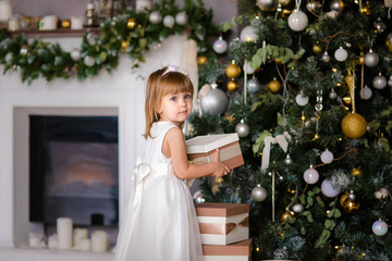 Cute little girl in the white dress with big presents near the Christmas tree