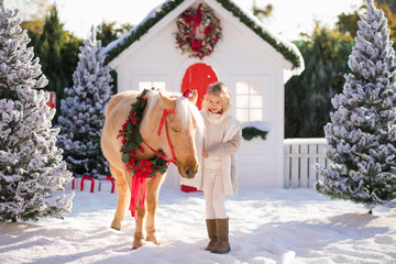 Nice blonde curly child caresses adorable pony with festive wreath near the small wooden house and...