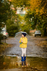 Child girl in yellow jacket and blue boots with umbrella runs through the autumn puddle