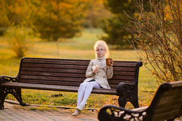 Curly schoolgirl in the glasses and light coat sitting on the branch in the autumn park and eating tasty cake and drinking tea on the sunset