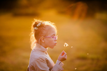 Curly schoolgirl in the glasses and light coat playing with dandelion in the autumn park on the sunset
