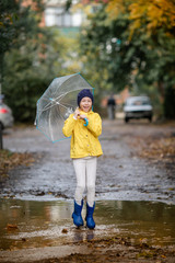 Child girl in yellow jacket and blue rubber boots with umbrella jumps in puddle on an autumn walk