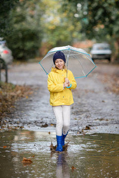 Child girl in yellow jacket and blue rubber boots with umbrella in puddle on an autumn walk