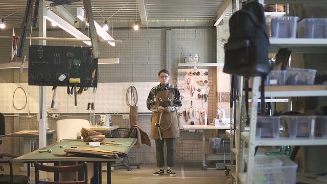 American young entrepreneur poses in workspace of modern leather workshop, front view of businessman is holding new real product for import, standing in lighting room, worker wearing stylish uniform