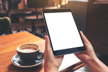 Mockup image of hands holding black tablet pc with blank white screen with coffee cup on wooden table in cafe
