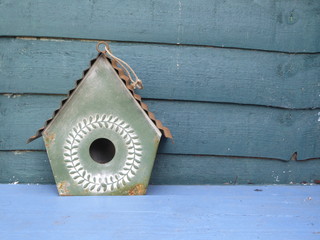 A green metal bird house on a blue wooden garden bench with a teal green fence 