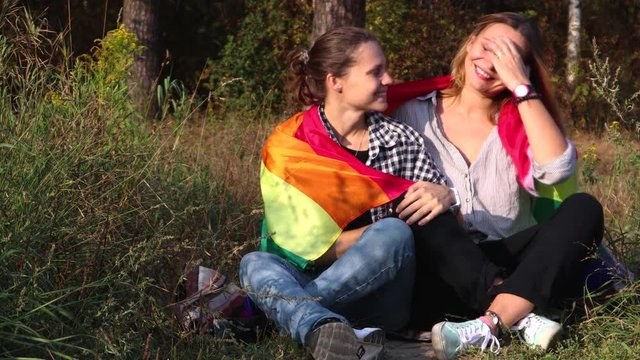 Two young women are sitting on a background of the rainbow flag. The sun is shining brightly, LGBT rights, lesbian family.