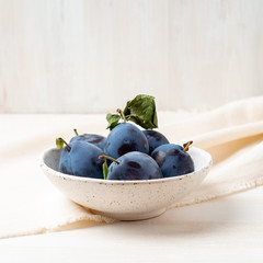 violet blue ripe plums in white bowl on white wooden table with linen textile ranner tablecloth, side view