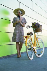 Naklejka premium pretty girl with mobile phone near wall and bicycle in city in summer