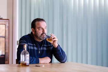 Senior man sitting at the dining room table having a glass of whiskey with the nearly empty bottle beside him