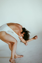 A slim girl with curly hair is moving, dancing, unusually posing with a view on her beautiful open back dressed in a white bodysuits on a white background