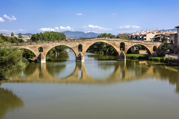 Fototapeta na wymiar Spain, Puente La Reina, Gares: Panorama view of famous romanesque bridge over river Arga with skyline of Spanish small town literally named 'Bridge of the Queen', green riverside and blue sky.