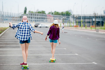 Two blond girls wearing checkered shirts, caps and denim shorts are longboarding on the empty car park. Sport and cool style.